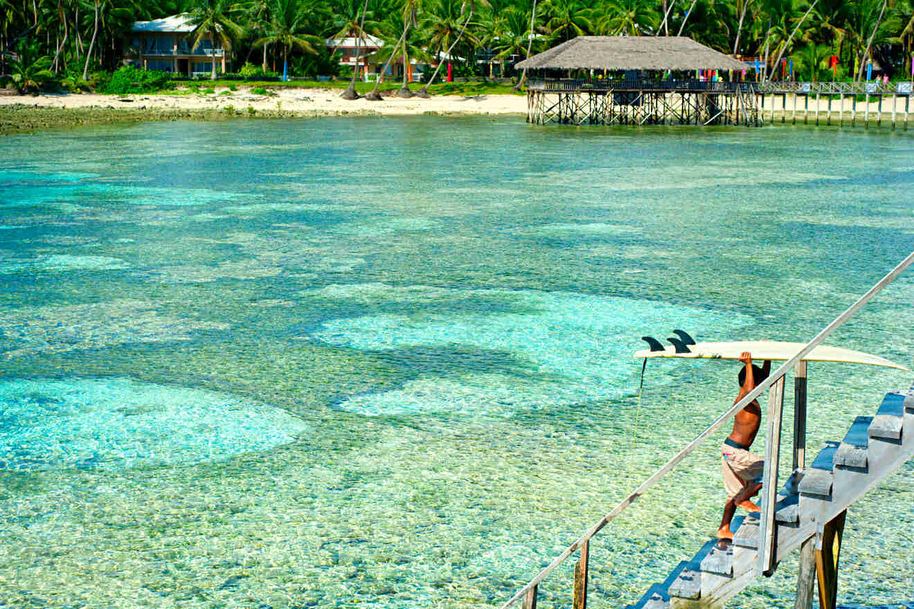 A man carrying a surfing board on a wooden pier overlooking crystal-clear waters with vibrant coral visible beneath, and traditional huts along the shore.